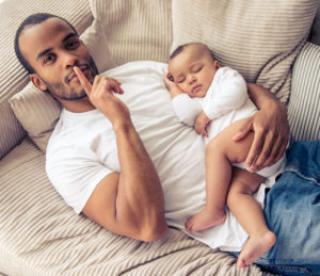 high angle view of handsome young afro american father looking at camera and showing silence sign while lying on sofa at home. little baby is sleeping in dad's arms 